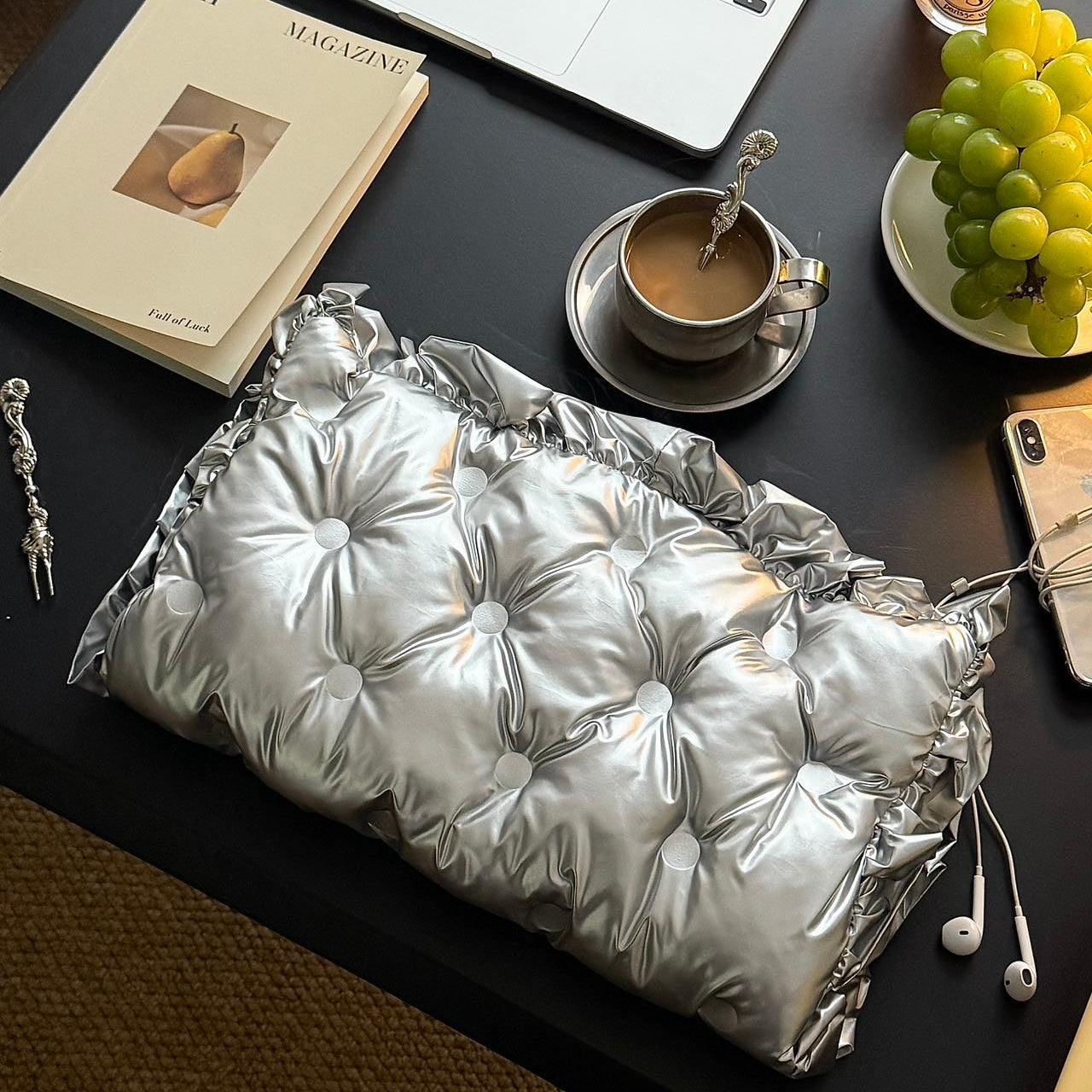 Silver Ruffle Graphene Hand Warmer | Portable USB Heating Bag displayed on a stylish workspace with coffee, magazine, and fresh grapes, featuring a quilted metallic silver finish, perfect for cozy desk use, home comfort, and portable warmth in everyday settings.
